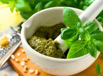 On the table there is a white bowl full of green pestle and fresh glossy leaves of the best basil for pesto among the ingredients and kitchen utensils.
