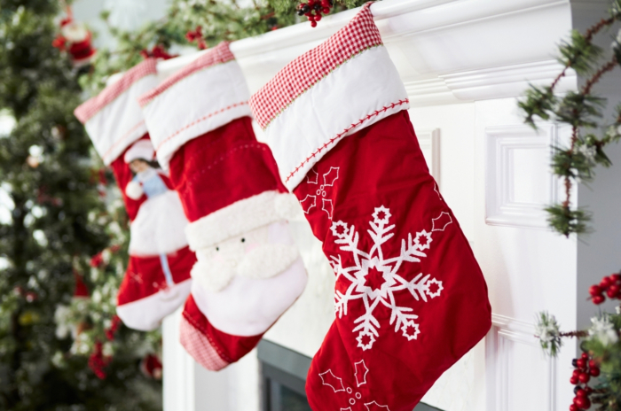 Three red Christmas stockings featuring images of a snowflake, Santa Claus, and a girl in a white dress hang on a decorated white fireplace, next to an indoor Christmas tree, perfect for filling with gardening stocking stuffers.