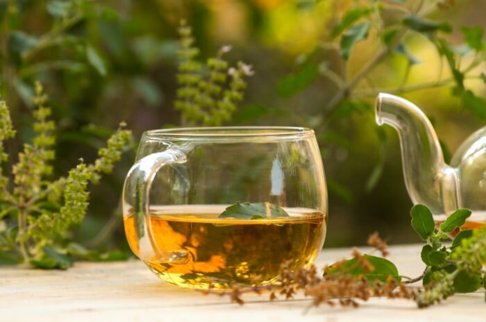 A clear glass mug sitting on an outdoor table displays golden tea and an herbal garnish next to a silver teapot.