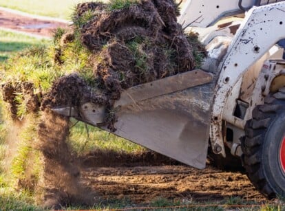 A view of a small bulldozer removing grass from a yard. The bulldozer appears worn and is white with thick rubber wheels and red rims. The surrounding green grass is still attached and rooted in dark brown soil. Clumps of earth can be seen being picked up by the bulldozer for pool installation preparation.
