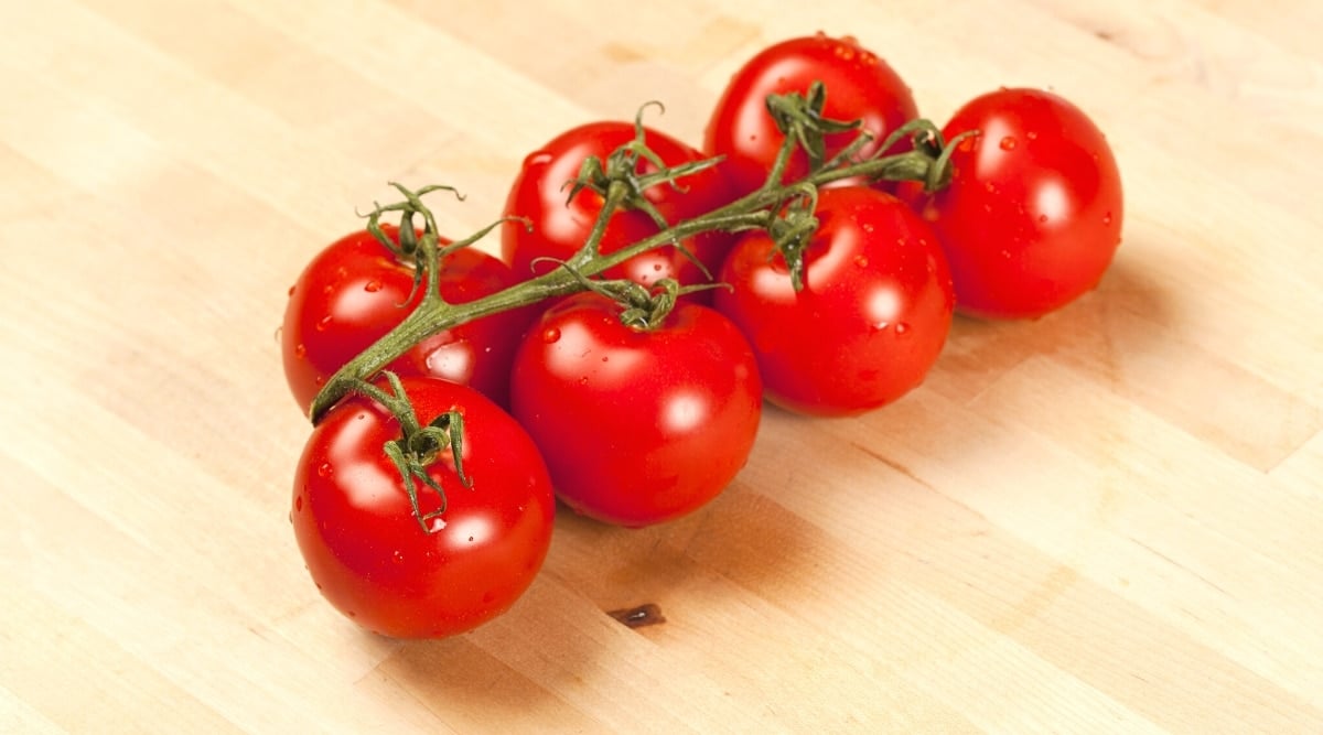 Image showing a bunch of seven round bright red fruits with water droplets growing on a vine placed on a light brown wooden surface. The fruits have the green stem still on them and some green creased leaves.